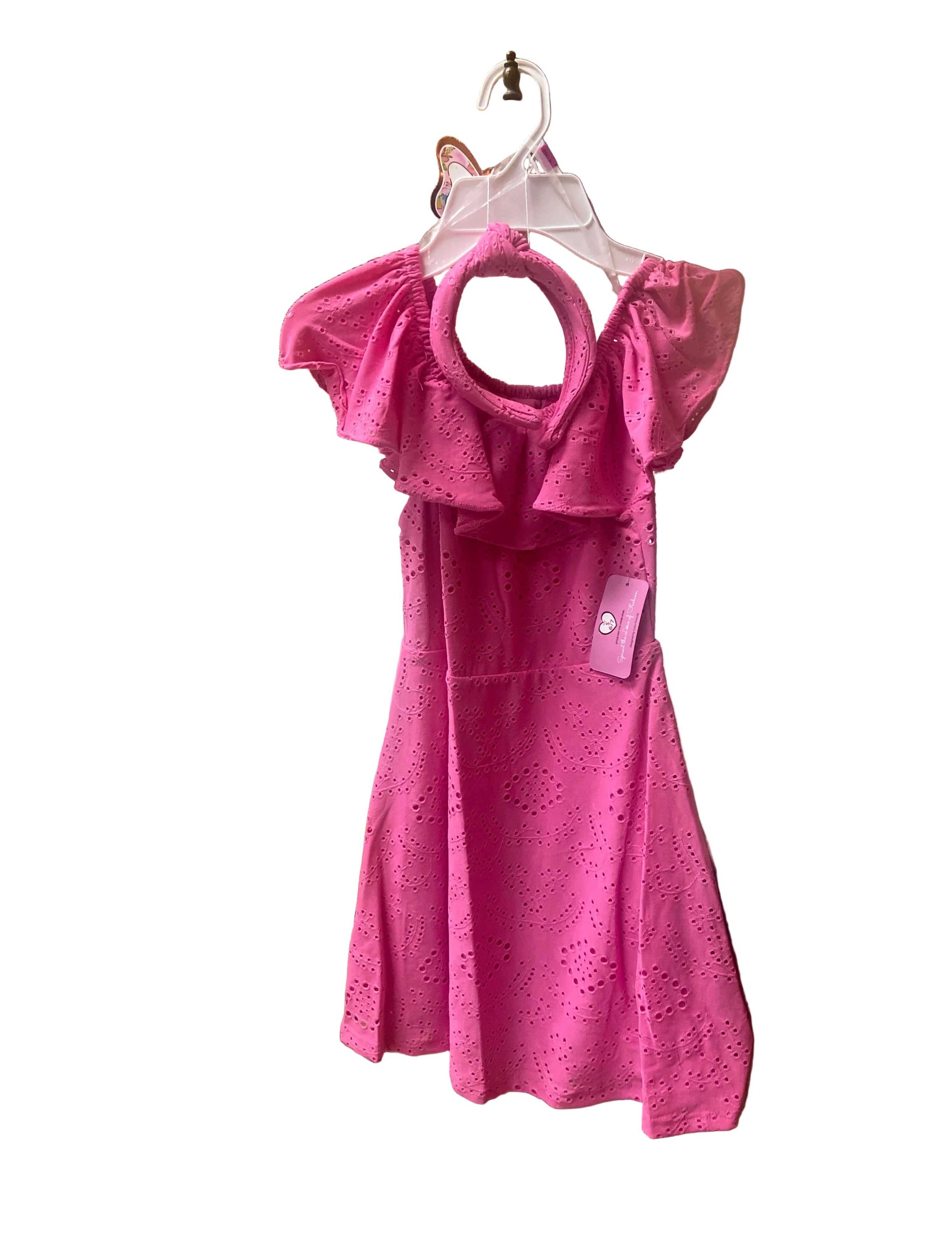 The Eyelet Dress is a bright pink, off-shoulder dress with ruffled sleeves and eyelet details, hanging on a white hanger with a matching pink headband. The background is white.