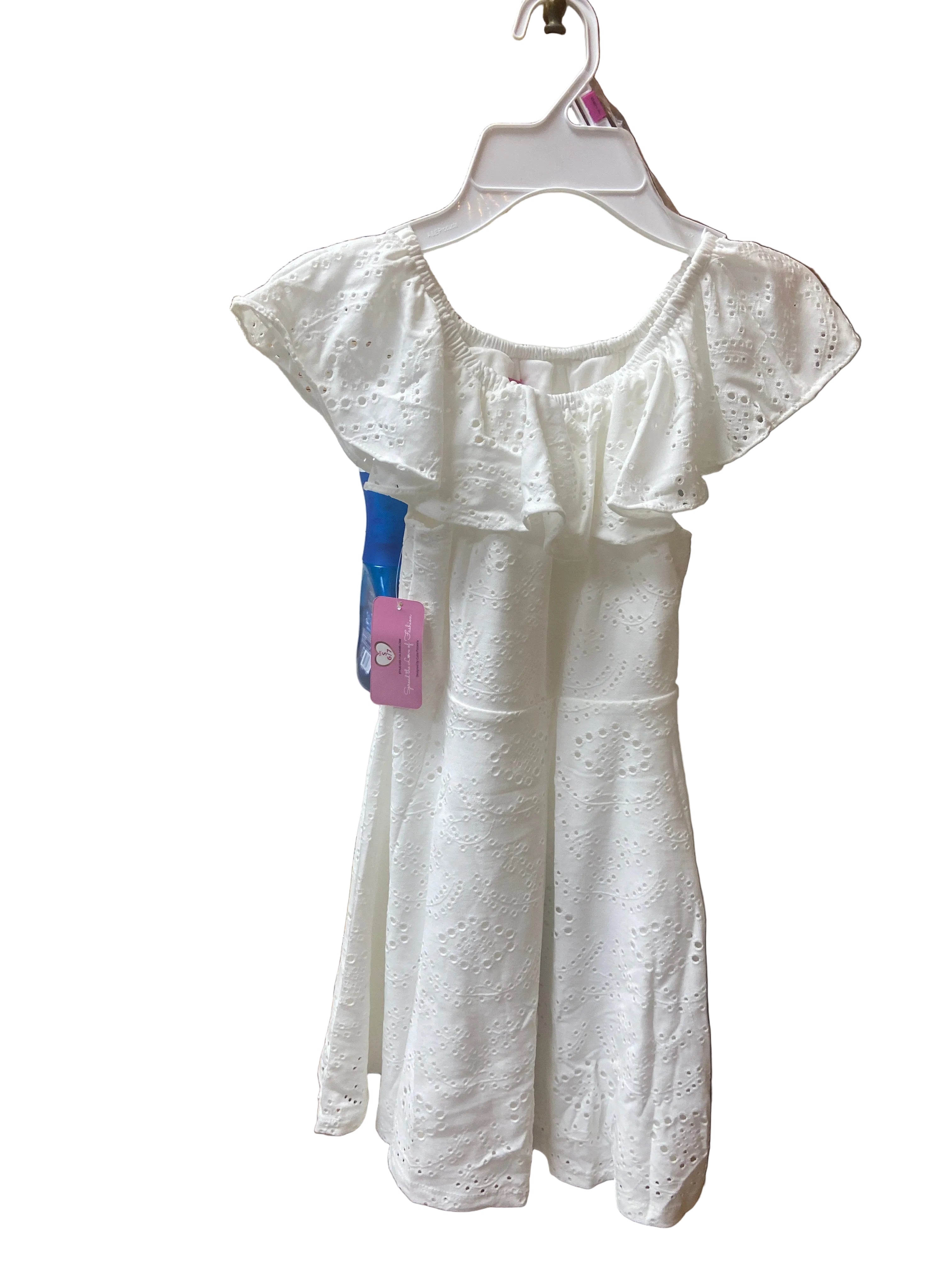 The Eyelet Dress with ruffled sleeves and a pink tag hangs on a hanger in a kitchen, featuring wooden cabinets, a sink, and a toaster in the background. A blue toothbrush is also attached to the dress.
