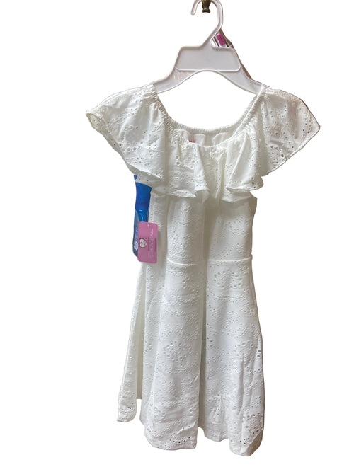 The Eyelet Dress with ruffled sleeves and a pink tag hangs on a hanger in a kitchen, featuring wooden cabinets, a sink, and a toaster in the background. A blue toothbrush is also attached to the dress.